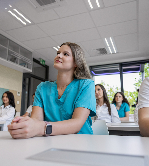 Žena ve zdravotnickém uniformě sedí v učebně s dalšími lidmi, poslouchá pozorně.