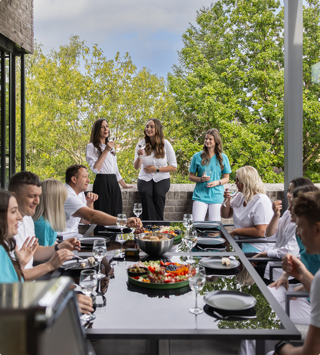 Skupina lidí v lékařských uniformách sedí u stolu s nádobím a předkrmy, zatímco tři ženy stojí a připíjejí si sklenicemi venku na terase za slunečného dne.