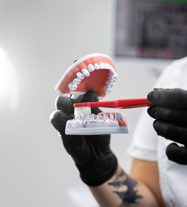 Osoba v černých rukavicích drží zubní model a ukazuje čištění zubů červeným zubním kartáčkem.