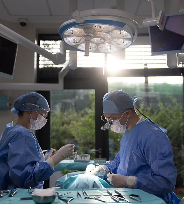 Dva chirurgové v modrém operačním oblečení a rouškách provádějí chirurgický zákrok v operačním sále s nástroji na stole.