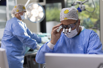 Chirurg v modrém ochranném oděvu upravuje zvětšovací brýle během operace v nemocnici.