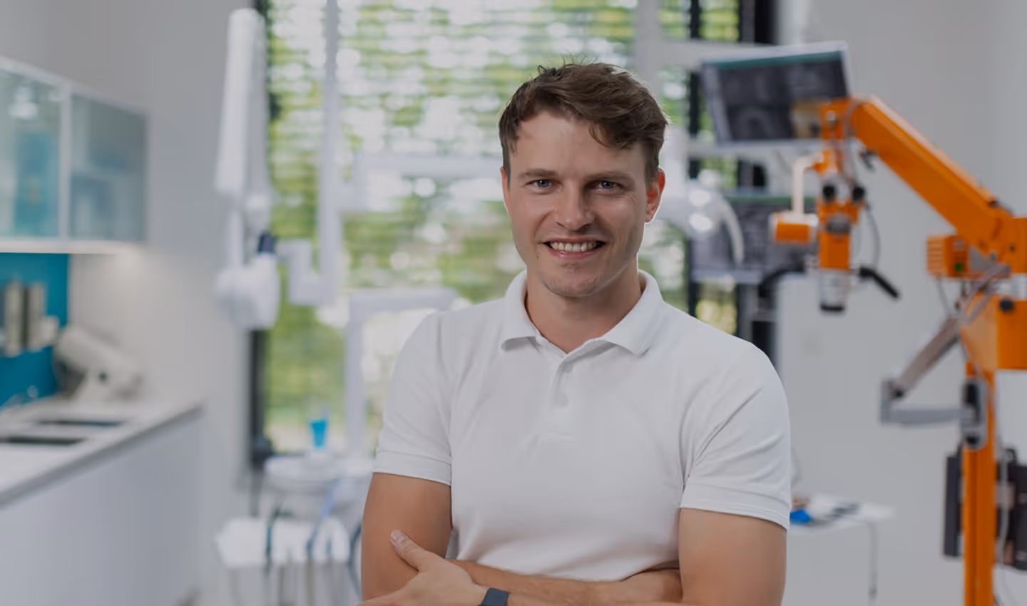 Usměvavý muž s bílým tričkem stojí v moderní zubní ordinaci s lékařským vybavením v pozadí.