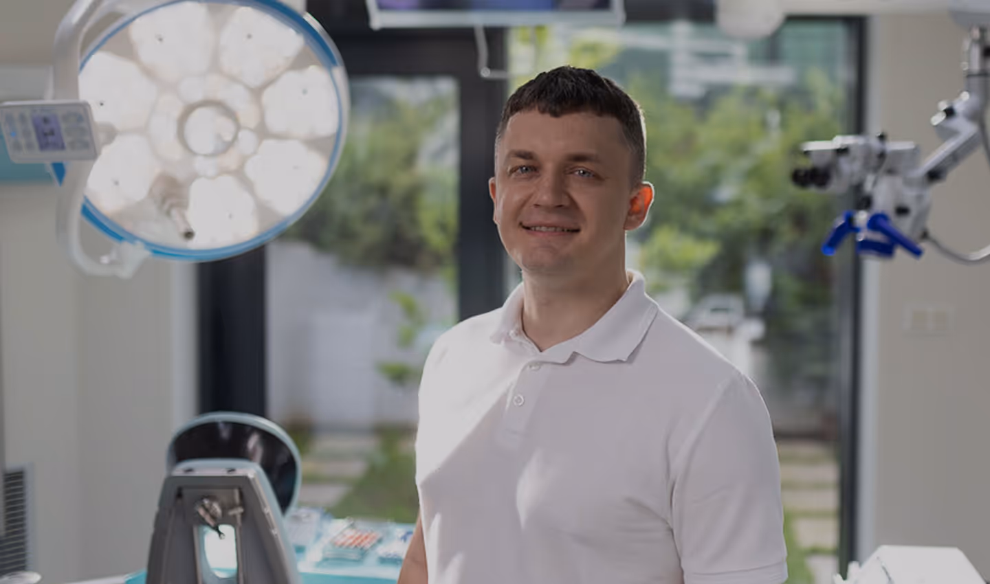 Muž v bílé polo košili stojící v zubní ordinaci s lékařským vybavením v pozadí.