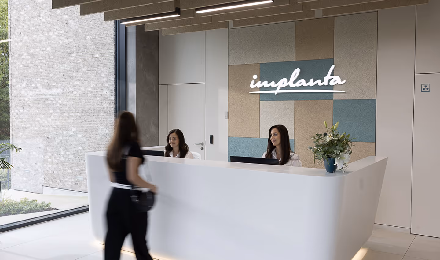 Recepce moderní kliniky s dvěma recepčními za bílým pultem a ženou přicházející k pultu, na stěně nápis 'implanta'.