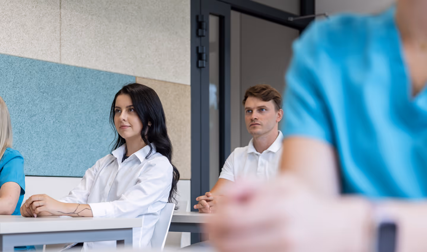 Dva mladí dospělí sedí v třídě a poslouchají, zatímco další osoba v modrém scrubu je v popředí rozmazaná.