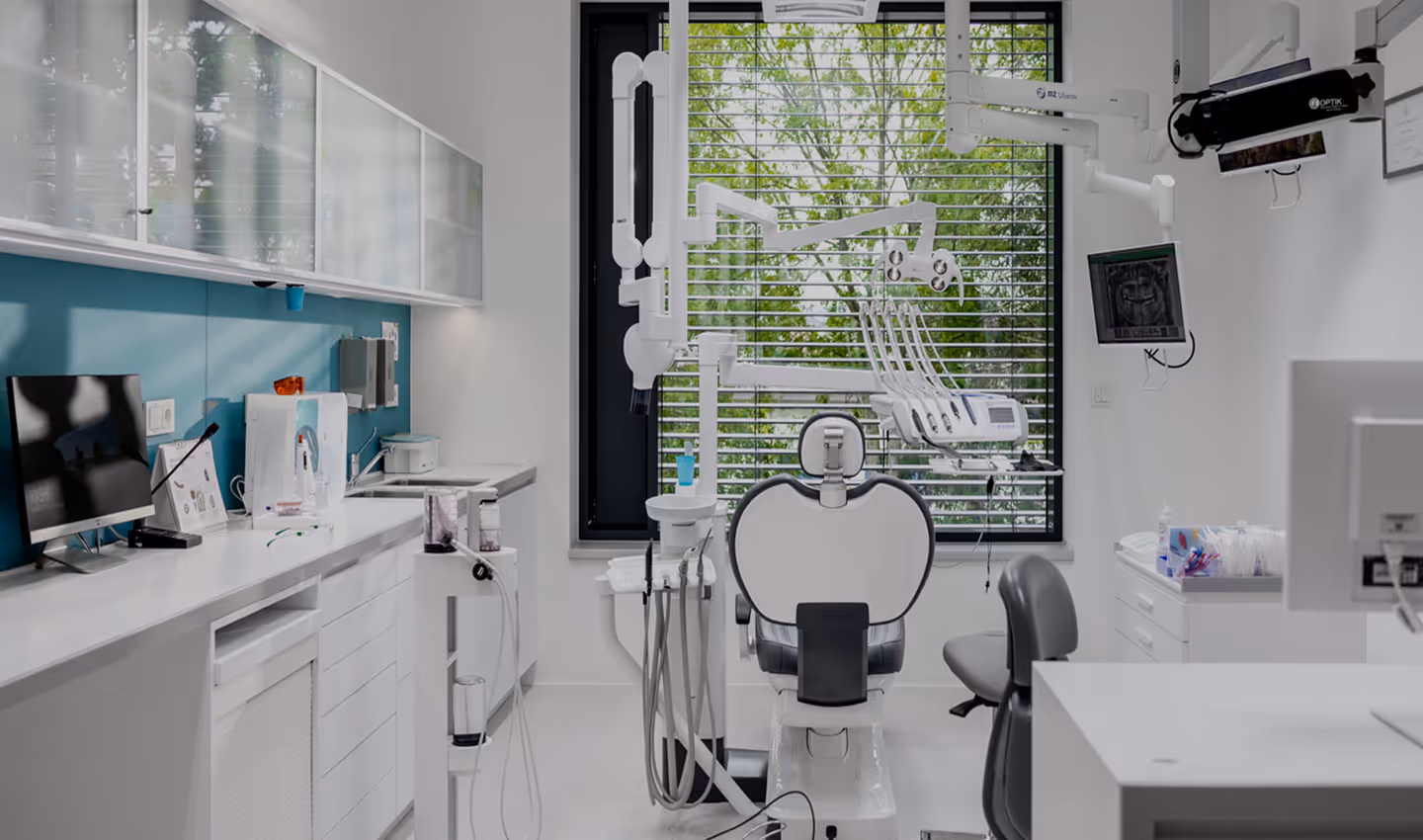 Moderní zubní ordinace s bílým stomatologickým křeslem uprostřed a zařízením na ošetření zubů, vedle velkého okna s žaluziemi.