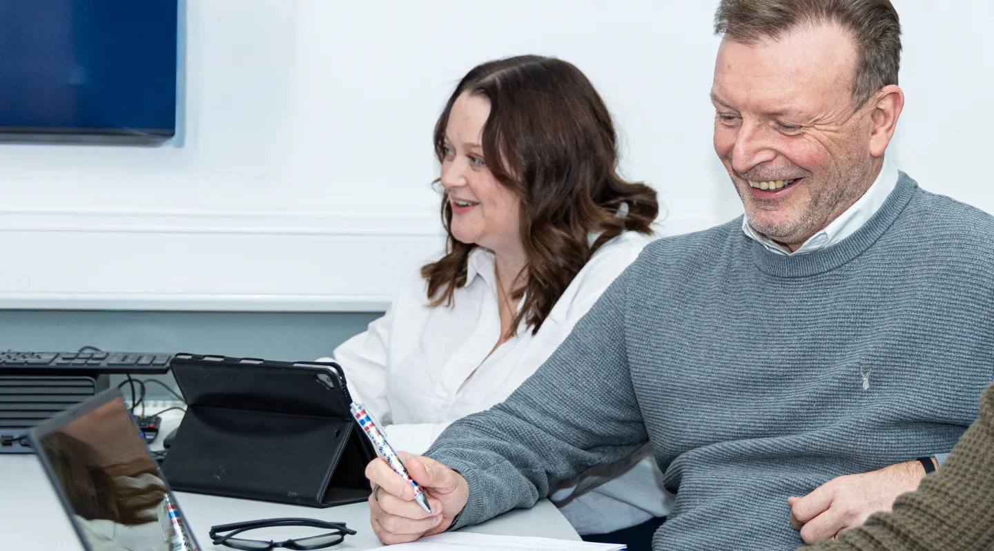 Two colleagues smiling and having a discussion in a meeting room with a tablet and laptop on the table.