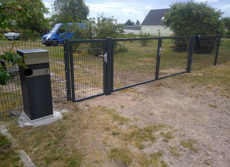 Schwarz lackiertes Gartentor mit Zaun und Briefkasten auf einem grassigen, teilweise sandigen Boden in einer Wohngegend.