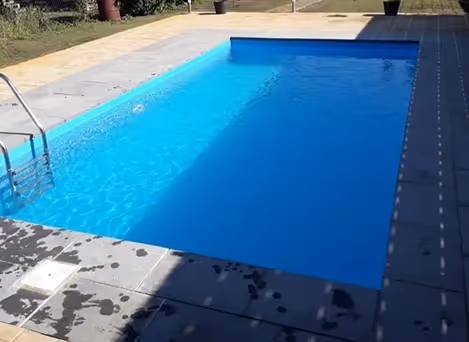 Ein klarer rechteckiger Schwimmbecken mit blauem Wasser und grauem Fliesenrand, teilweise von Schatten bedeckt.