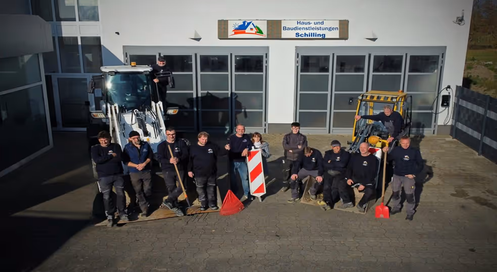 Gruppenfoto von zwölf Bauarbeitern vor einem Gebäude mit Maschinen und Werkzeugen, unter einem Schild mit der Aufschrift Haus- und Baudienstleistungen Schilling.