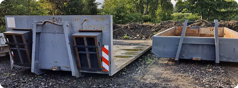 Zwei große, graue Metallplattformen auf unebenem Schottergelände, umgeben von Bäumen und Sträuchern.