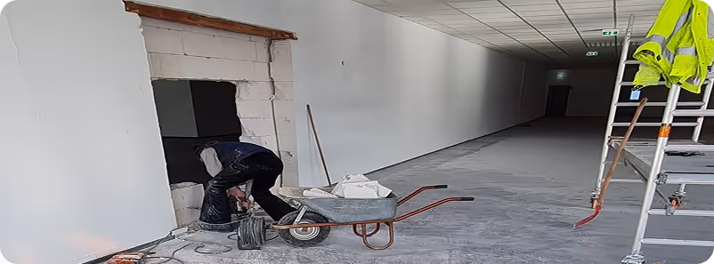 Bauarbeiter arbeitet in einem langen, leeren Gang neben einer Radschubkarre und einer Leiter mit gelber Schutzjacke.