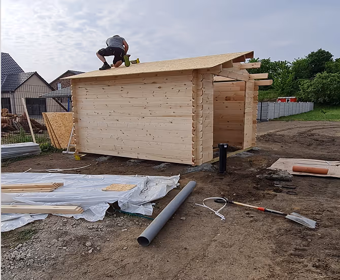 Person arbeitet auf dem Dach eines kleinen, aus Holz gebauten Gartenhauses auf einer Baustelle.