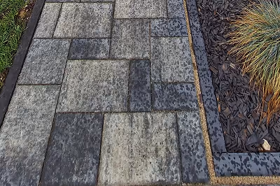 Nasse, rechteckige graue Steinfliesen als Gehweg neben einem Beet mit Mulch und Gräsern.