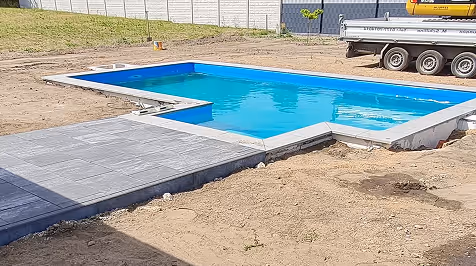 Partially installed backyard swimming pool with blue water and surrounding unfinished ground with a paved area on one side.
