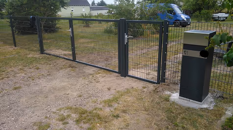 Metallzaun mit einem Tor und einer schwarzen Briefkastenstation neben einem grasbewachsenen Weg.