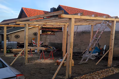 Holzkonstruktionsrahmen für eine überdachte Terrasse entsteht vor einem Haus mit rotem Ziegeldach.