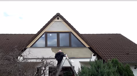 Person auf einer Leiter streicht die Fassade eines Hauses mit einem dreieckigen Giebel.