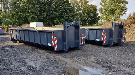 Zwei leere graue Absetzmulden stehen auf einem Kiesplatz vor Bäumen an einem bewölkten Tag.