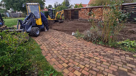 Gartenarbeiter an einer kleinen Radlader-Maschine auf einem gepflasterten Weg neben einem frisch umgegrabenen Boden unter Sträuchern und Bäumen.