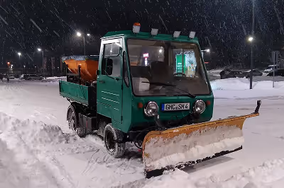 Kleiner grüner Schneeräumfahrzeug mit Schneepflug bei Schneefall auf schneebedecktem Parkplatz.