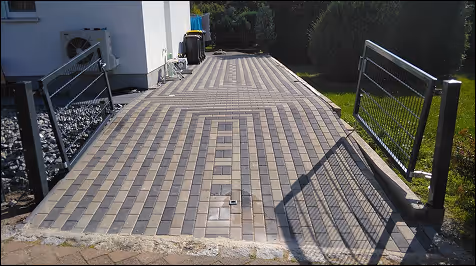 Einfahrt mit grau-gelben rechteckigen Pflastersteinen, links ein Stahlzaun und rechts ein offenes Tor mit Rasenfläche dahinter.