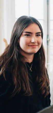 Jeune femme aux longs cheveux bruns souriant légèrement devant une fenêtre lumineuse.