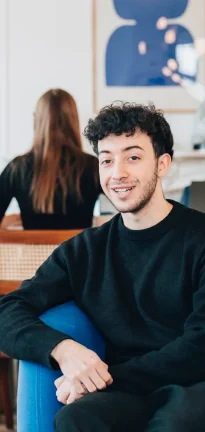 Jeune homme souriant en pull noir assis sur une chaise bleue, avec une femme aux longs cheveux bruns de dos en arrière-plan.