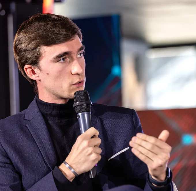 Homme portant un costume bleu foncé et un col roulé noir, tenant un microphone et s'exprimant lors d'une discussion.