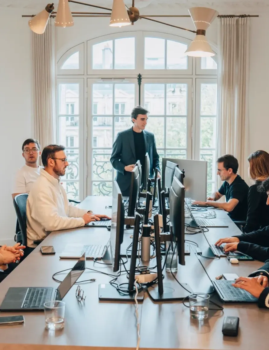 Un groupe de personnes travaille autour d'une grande table de conférence avec plusieurs écrans d'ordinateur dans un bureau lumineux avec une grande fenêtre en arc.