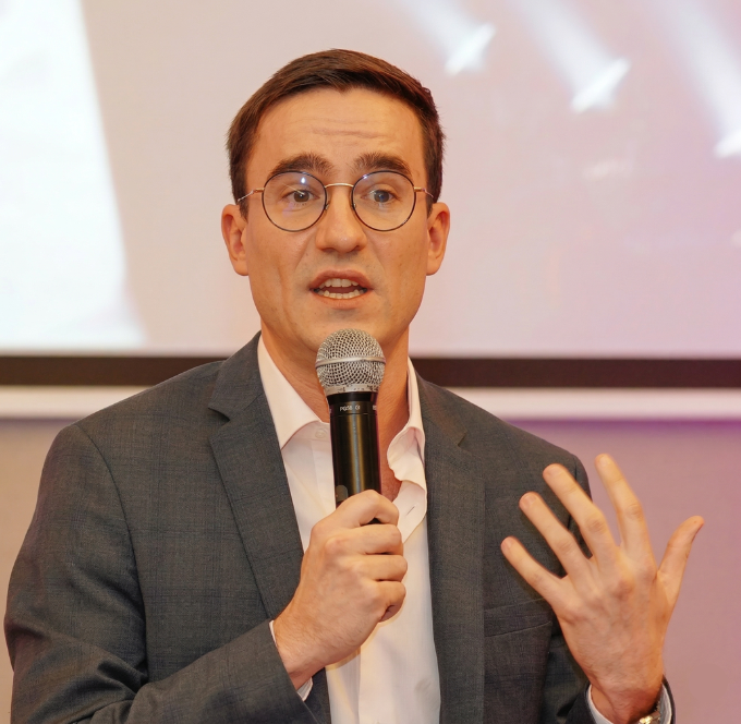 Un homme parlant dans un micro lors d'un événement, portant un blazer bleu et un col roulé noir.