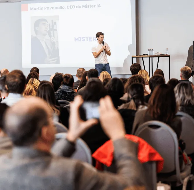 Homme parlant avec un micro lors d'un événement ou d'une conférence, habillé en veste bleu foncé et col roulé noir.