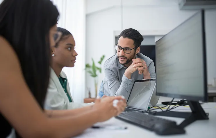 Trois personnes en réunion dans un bureau, regardant un grand écran d'ordinateur sur un bureau.