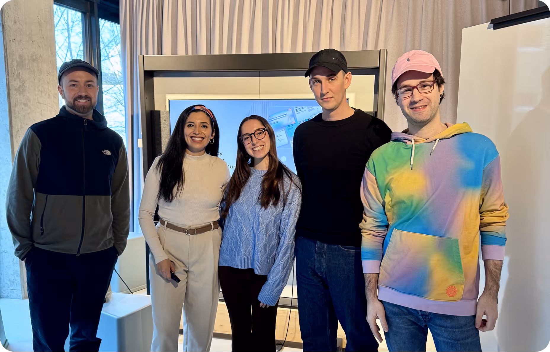 Five casually dressed people standing indoors smiling in front of a large screen and window.