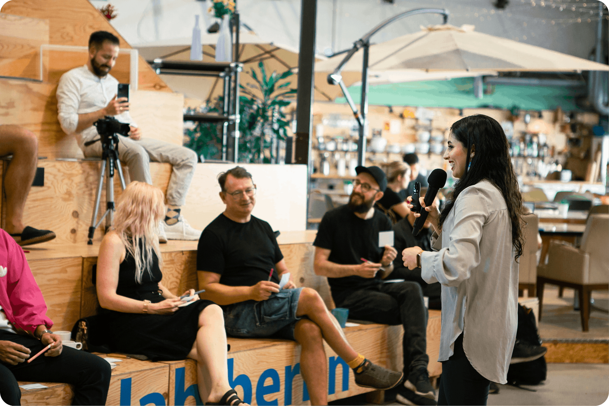 Woman speaking into a microphone while an audience seated on wooden steps listens and interacts in a casual indoor event space. Gaby workshop Motion Lab