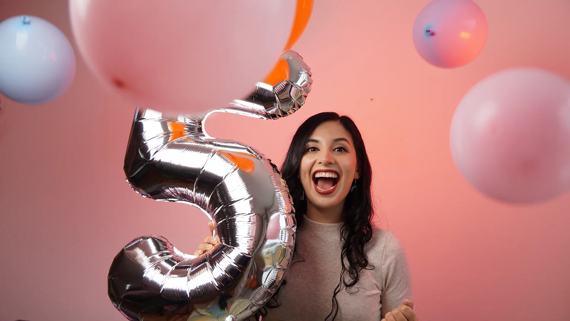 Smiling woman holding a shiny silver balloon shaped like the number five surrounded by floating balloons.