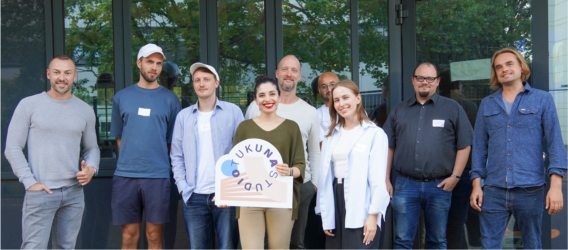 Group of nine people standing in front of glass doors, smiling, with one woman who is Gaby Corral holding a sign that reads 'TUKUNA STUDIO'. Workshop in BHT