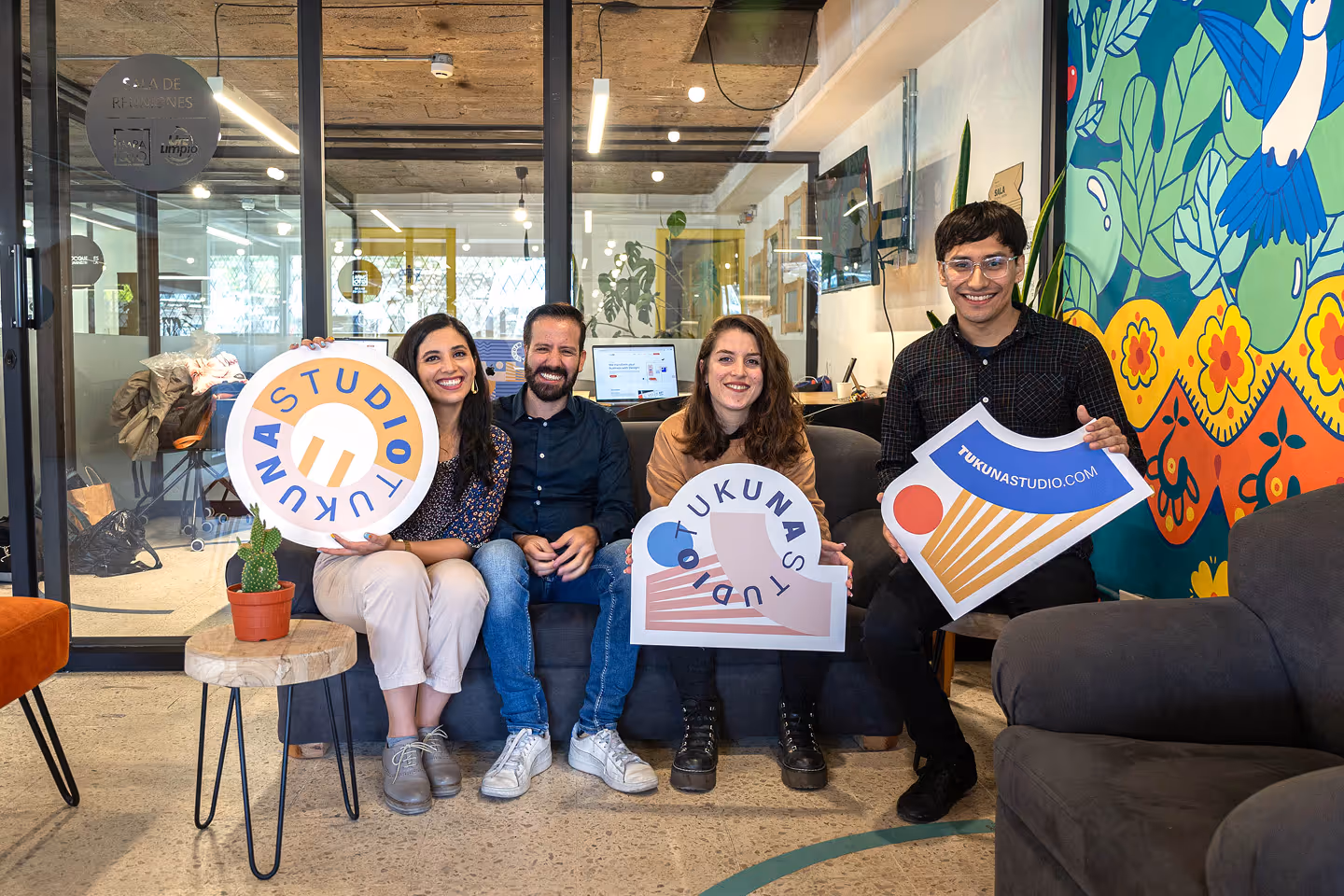 Four people sitting on a couch in a colorful office holding Tukuna Studio logo signs.