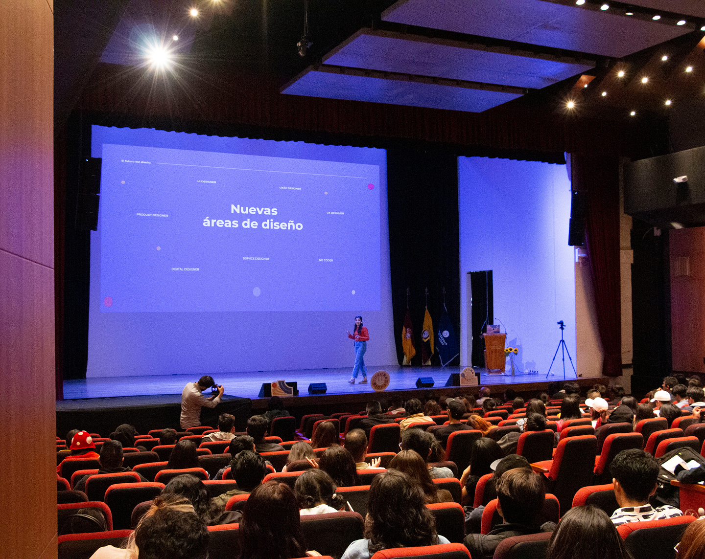 Speaker presenting on stage in an auditorium with a slide titled 'Nuevas áreas de diseño' and an audience seated in red chairs. Gaby talk in UDA