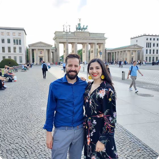 A smiling couple stands in front of the Brandenburg Gate in Berlin on a bright day.