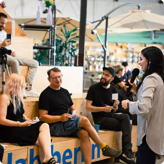 Woman speaking into a microphone to seated people taking notes in a casual indoor setting.