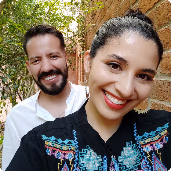 Smiling woman with embroidered blouse and man with beard standing outdoors near a brick wall and greenery.