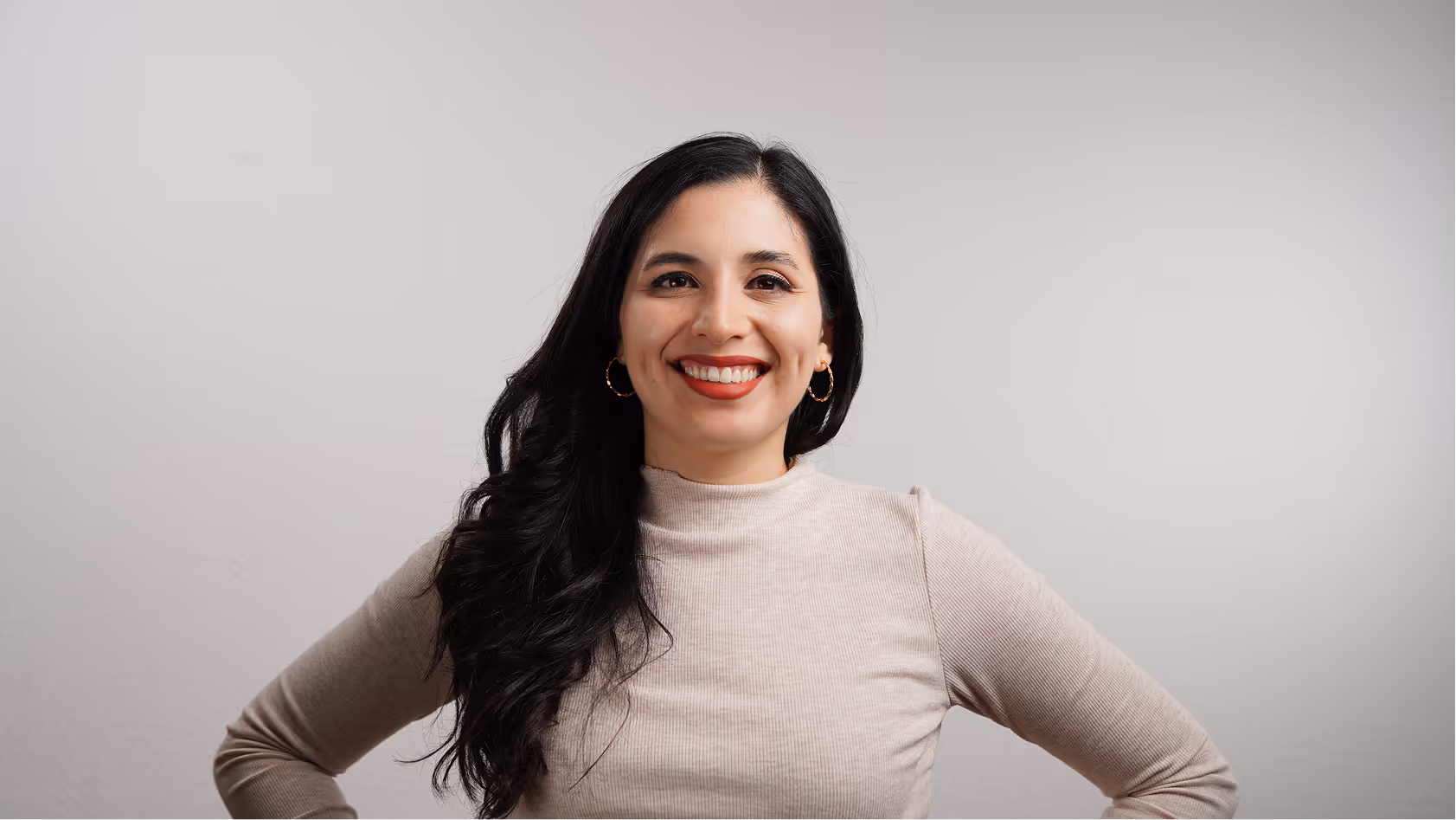Smiling woman with long black hair wearing a beige turtleneck sweater and hoop earrings standing against a plain gray background.