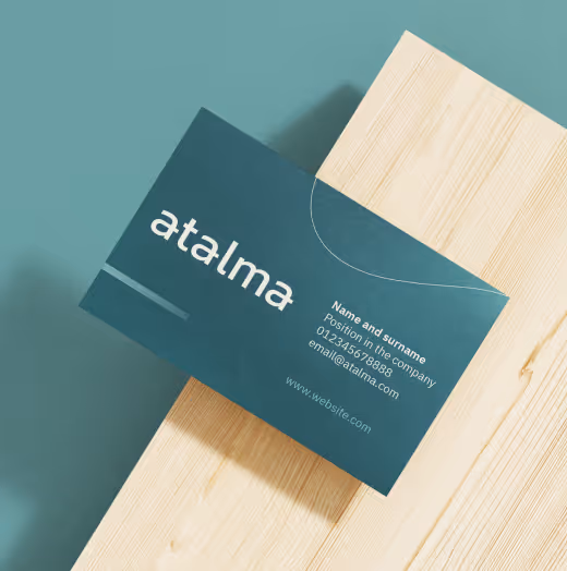 Dark teal business card with white text showing company name 'atalma' and placeholder contact details on a wooden surface.