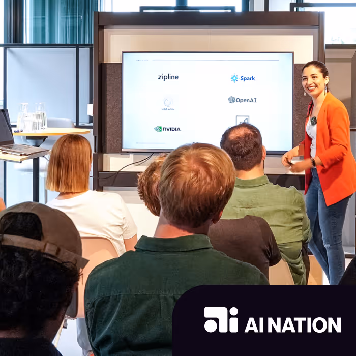 A woman in an orange blazer presenting to a seated audience, with a screen displaying logos including zipline, Spark, OpenAI, Miso Acua, NVIDIA, and AI Nation.
