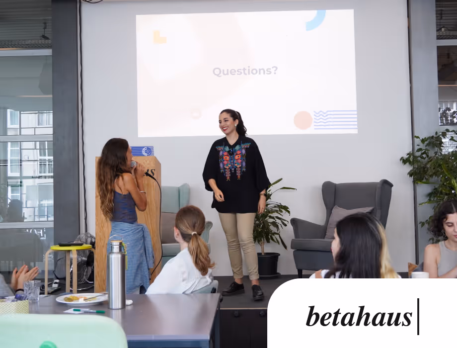 Two women on stage during a presentation, one speaking into a microphone, with an audience seated and a screen behind displaying the word 'Questions?'.