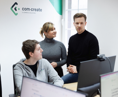 Three coworkers collaborating in an office with computers, under a com-create e-commerce logo.