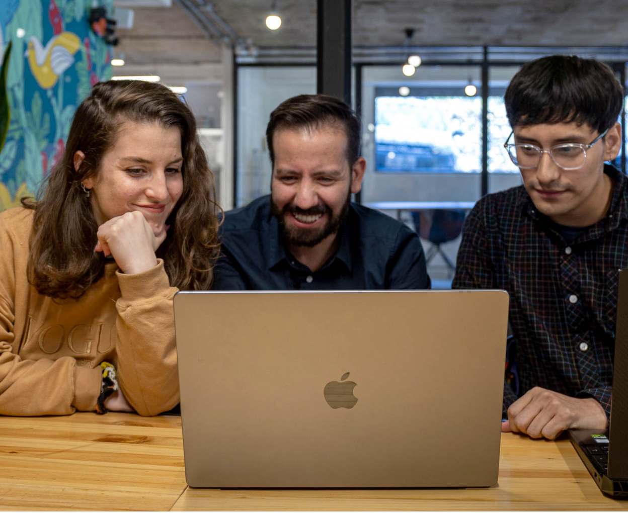 Three colleagues sitting at a wooden table, smiling and looking at a laptop screen in a modern office setting.