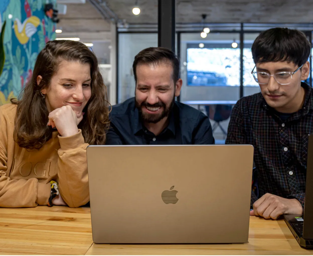 Three colleagues sitting at a wooden table, smiling and looking at a laptop screen in a modern office setting.