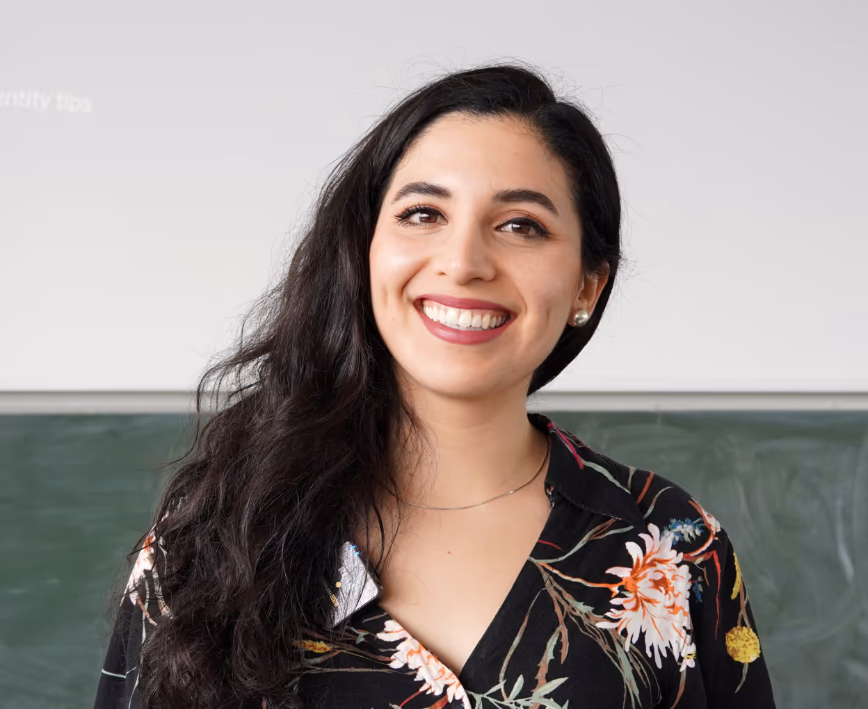 Smiling woman with long black hair wearing a black floral blouse standing in front of a chalkboard.